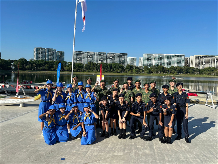 Group of youth in blue scout and dark navy uniforms posing outdoors near a waterfront with buildings in background.